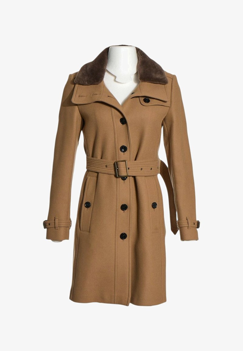 Long manteau camel pour femme avec col en fourrure foncée, boutons noirs sur le devant, taille ceinturée et poignets à boucles.