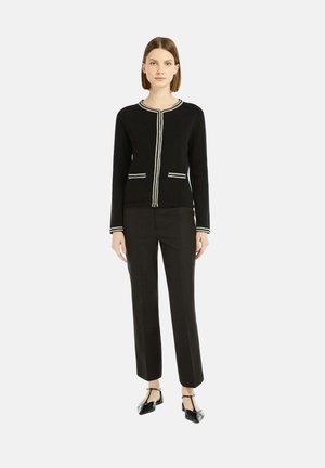 Vrouw staat, draagt een zwarte lange mouwen vest met witte biezen, zwarte rechte broeken en zwarte T-band schoenen tegen een witte achtergrond.