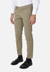 Chinos beige a gamba affusolata con una texture liscia, tasca frontale e chiusura a un bottone, indossati con scarpe nere e calzini.