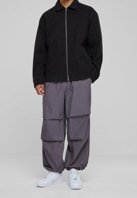 Svart zip-up jacka med krage, tillsammans med lösa grå cargobyxor med flera veck och vita sneakers.