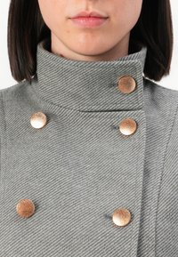 Detailní záběr osoby v šedém vlněném kabátu s vysokým límcem a pěti lesklými měděnými knoflíky uspořádanými asymetricky.