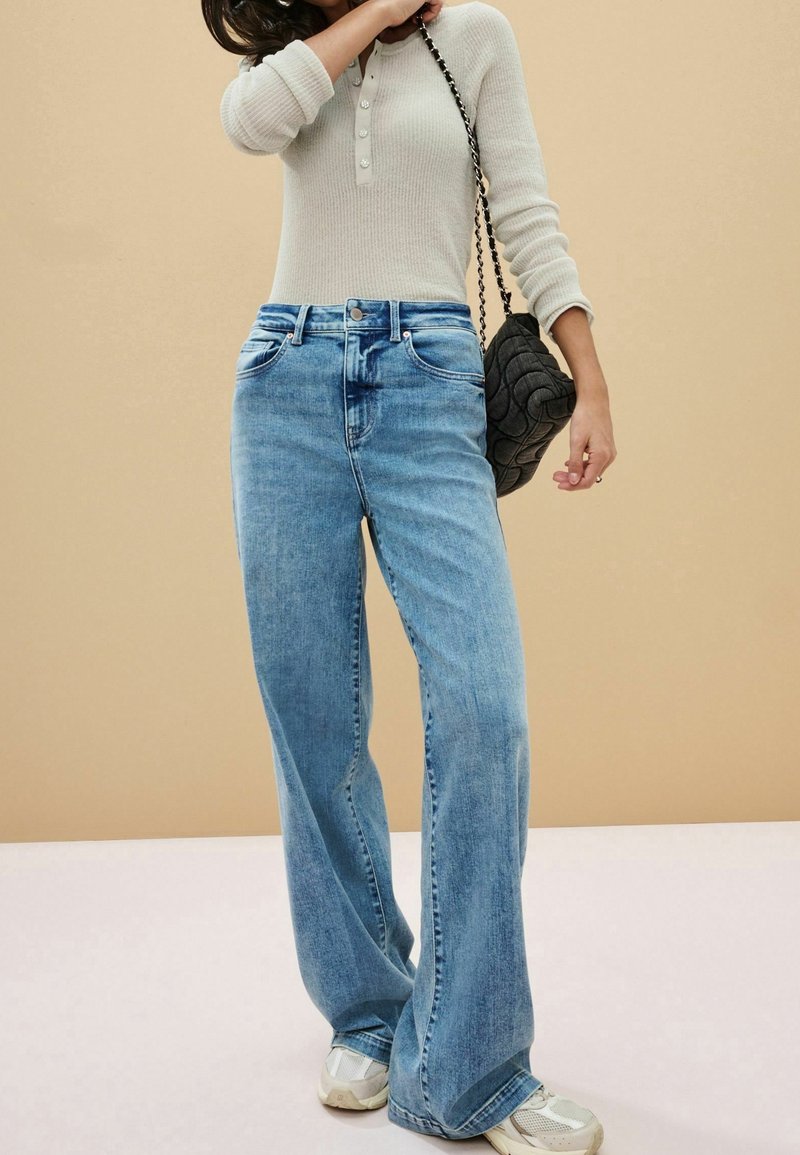 Lichtblauwe wijde jeans met hoge taille en vijf-pocket ontwerp, gecombineerd met een textuurbeige lange mouwen top en een zwarte gewatteerde tas.