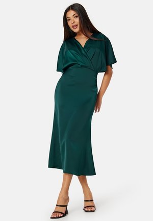 Robe en satin vert émeraude avec un design enroulé, manches courtes évasées et silhouette ajustée, atteignant le mi-mollet. Portée avec des talons noirs à brides.