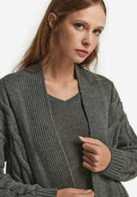 Grå stickad cardigan med ribbad krage och texturerad mönster. Lager på en matchande grå v-ringad topp med en mjuk textur.