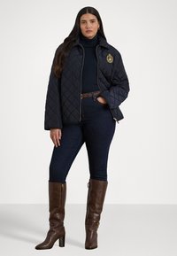 Quiltet marineblå jakke med høy krage og gyldent logo, sammen med en svart turtleneck, mørk denimjeans og brune kne-høye støvler.