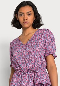Vestido floral cor-de-rosa com mangas bufantes, decote em V, cintura ajustada e barra franzida. Apresenta um padrão de rosas repetido em tons de vermelho e azul.