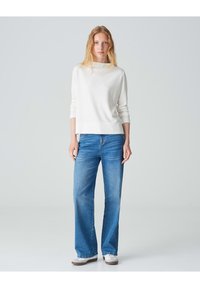 Weißer gerippter Pullover mit hohem Kragen, kombiniert mit blauen weiten Jeans. Das Outfit wird durch hellfarbene Sneaker ergänzt.