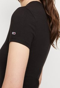 Černé tričko s krátkým rukávem vyrobené z bavlny. Na levém rukávu je malé logo s červenými a bílými akcenty. Hladká textura.