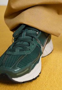 Sapatilhas de malha verde com detalhes cinza, apresentando atacadores texturizados, um bico moldado e uma sola de amortecimento branca, combinadas com calças bege.