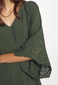 Olivgrünes Kleid mit V-Ausschnitt und kunstvoll mit Spitze besetzten Glockenärmeln. Weicher Stoff mit fließendem Fall und detaillierter Stickerei.