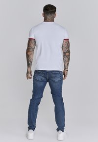 Witte T-shirt met korte mouwen en rode accenten, gecombineerd met blauwe denim jeans en witte sneakers, heeft een relaxte pasvorm en zichtbare tatoeëer-detail.