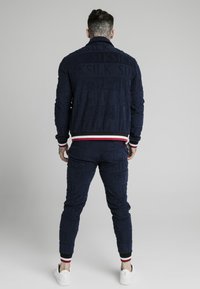 Navy tracksuit made of textured fabric, featuring an all-over logo pattern, contrasting striped cuffs, and white sneakers.