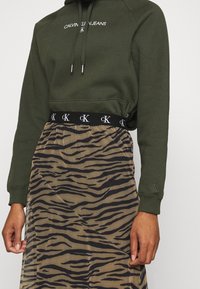 Olivgrön sweatshirt med logotyp, kombinerad med en genomskinlig kjol med ett mönster av svart och brunt tigerstripe och resårmidja med logotypdetalj.