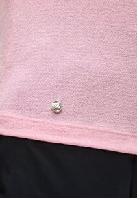 Ροζ υφασμάτινη υφή με μικρό στρογγυλό ασημένιο κουμπί που έχει σύμβολο καρδιάς και διασταυρωμένων οστών κοντά στην μαύρη κάτω άκρη του υφάσματος.