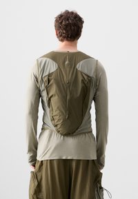Olivgrünes Langarmoberteil mit geschichtetem Stoffdesign, das Netzeinsätze und eine integrierte Reißverschlusstasche auf der Rückseite für Stauraum bietet.