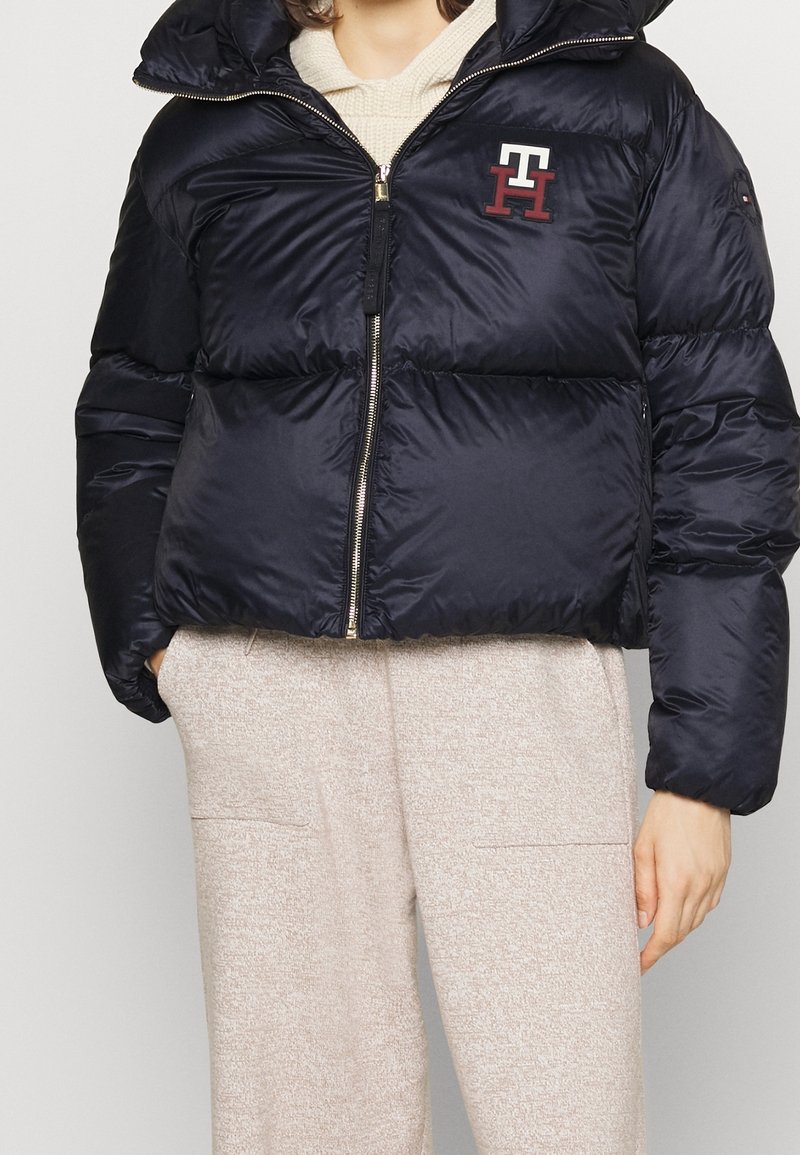 Marineblå jakke med dunfyld og glansfuld overflade, lynlås foran og hætte. Har et broderet maroon logo på brystet; cropped design.