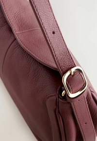 Gros plan sur une sangle de sac à main en cuir rouge foncé texturé avec une boucle dorée et un trou de réglage sur un fond uni.