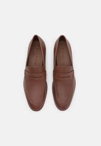 Mocassins en cuir marron avec une finition lisse, présentant un design surpiqué, une sangle décorative et un logo en relief sur la semelle intérieure.