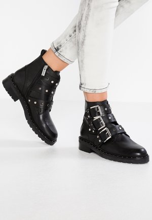 Personne portant des bottines en cuir noir avec trois sangles à boucle et des clous argentés, associées à un jean clair retroussé.