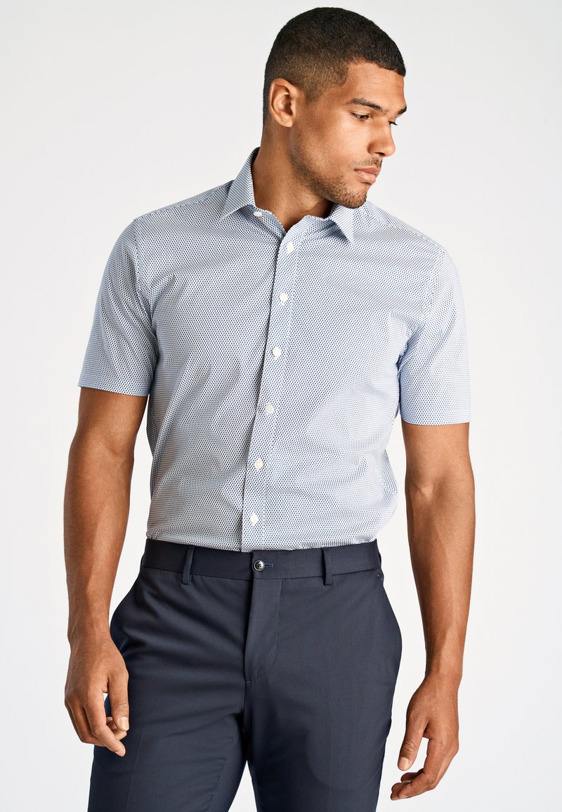 Camicia a maniche corte con bottoni di colore azzurro chiaro, adornata con un piccolo motivo geometrico bianco; abbinata a pantaloni blu scuro, con una texture di tessuto liscia.
