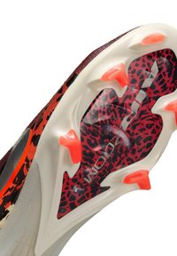 Gros plan de la semelle d'une chaussure de football rouge et noire à motif guépard avec une base blanche et des crampons orange, conçue pour terrain ferme.