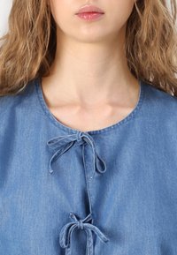 Top in denim azzurro chiaro con scollatura rotonda, dotato di due nastri in tessuto al centro davanti e una superficie leggermente texturizzata.
