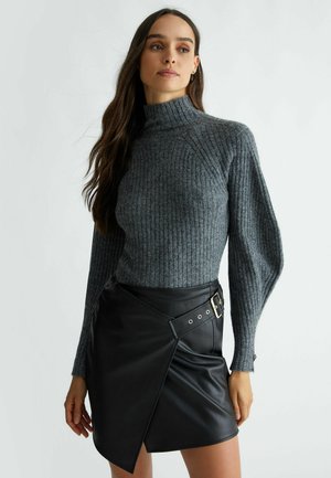 Pullover grigio a collo alto con maniche lunghe, abbinato a una mini gonna nera in ecopelle con design asimmetrico e dettaglio in fibbia metallica.