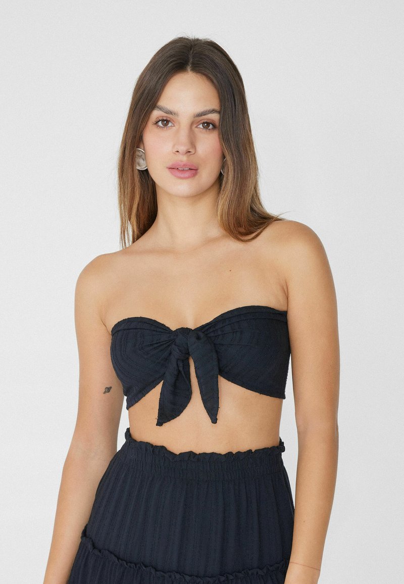 Czarny top bandeau z węzłowym detalem z przodu, wykonany z teksturowanej tkaniny i o dopasowanym kroju. Modelka nosi dopasowaną, warstwową spódnicę.