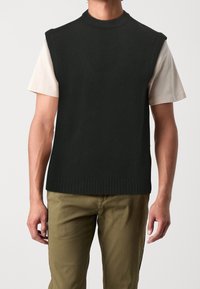 Man wearing a black sleeveless knit vest over a light beige short-sleeve t-shirt and olive green pants, standing against a plain background.