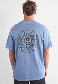 Maglietta a maniche corte azzurro chiaro con grafica circolare sul retro che presenta motivi neri e blu, e un piccolo etichetta sulla spalla sinistra.