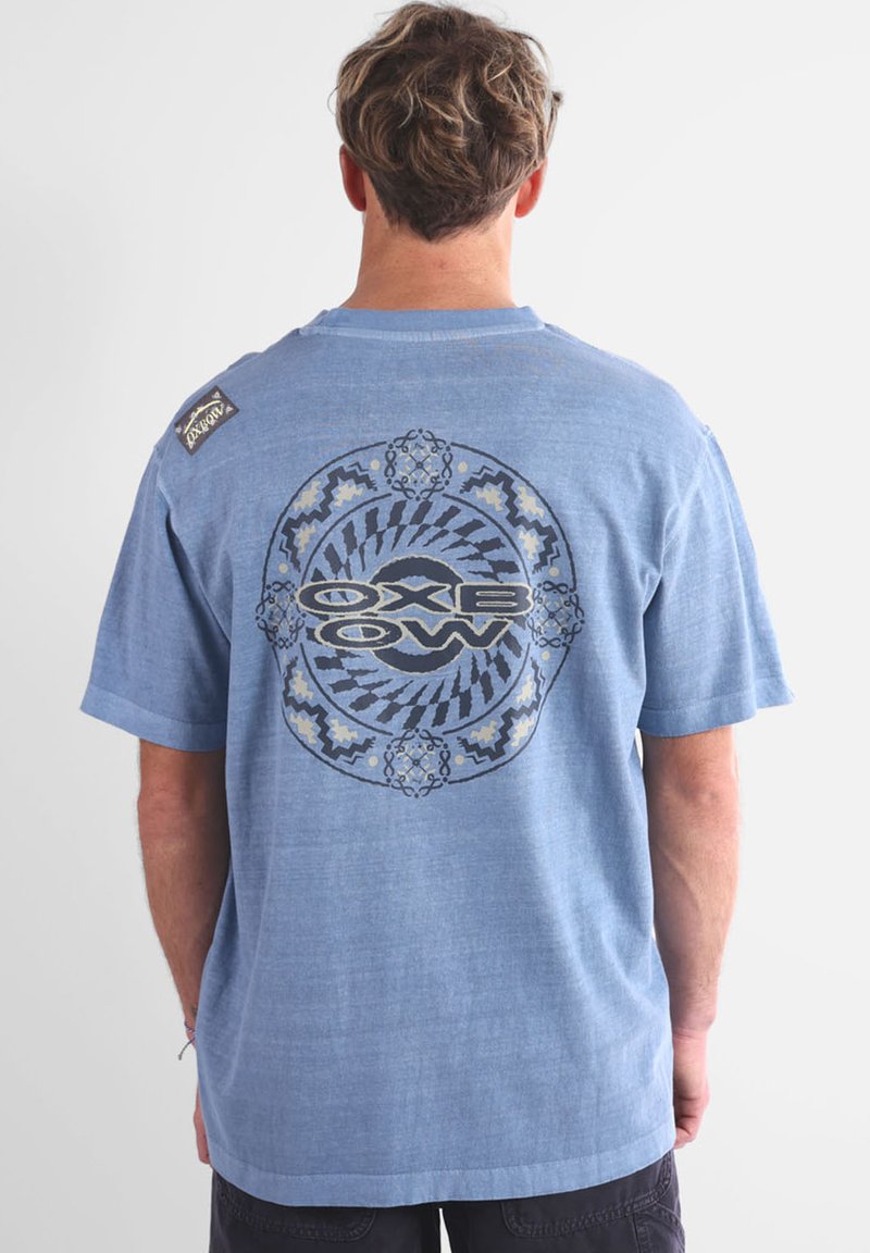 Maglietta a maniche corte azzurro chiaro con grafica circolare sul retro che presenta motivi neri e blu, e un piccolo etichetta sulla spalla sinistra.