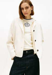 Roomkleurig, oversized jack met een zachte textuur, grote knopen en twee voorzakken, gedragen over een lichte grafische trui en donkere jeans.