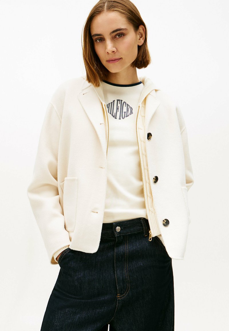 Roomkleurig, oversized jack met een zachte textuur, grote knopen en twee voorzakken, gedragen over een lichte grafische trui en donkere jeans.