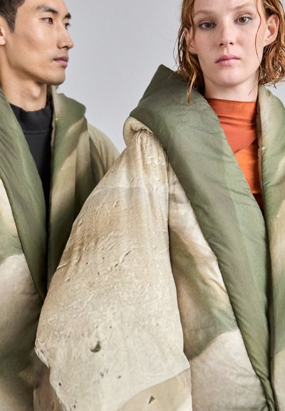 Oversized, textured outerwear in muted green and beige tones. Features a large hood and a flowing, draped shape with a layered fabric design.