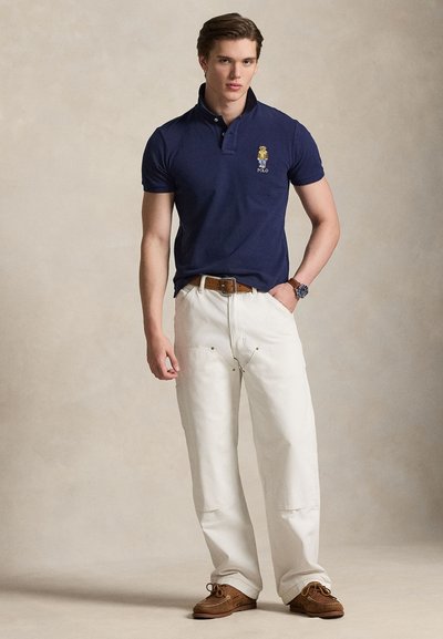 Polo bleu marine avec un logo, pantalon cargo blanc et chaussures bateau marron. La tenue présente une coupe classique et un tissu texturé.