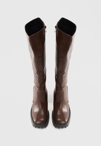 Bottes en cuir marron hautes avec des semelles en caoutchouc noir épaisses et des fermetures éclair sur le côté, se tenant droit sur un fond clair et uni.
