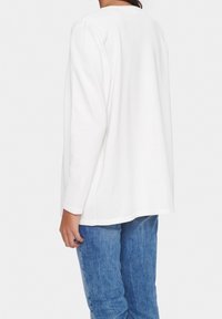 Kvinna som bär en vanlig vit långärmad skjorta och blå jeans, visad bakifrån mot en vit bakgrund.