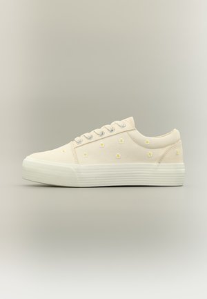Baskets basses de couleur crème avec semelle en caoutchouc blanche, œillets argentés, et petites marguerites brodées jaune et blanc sur la partie supérieure en tissu.