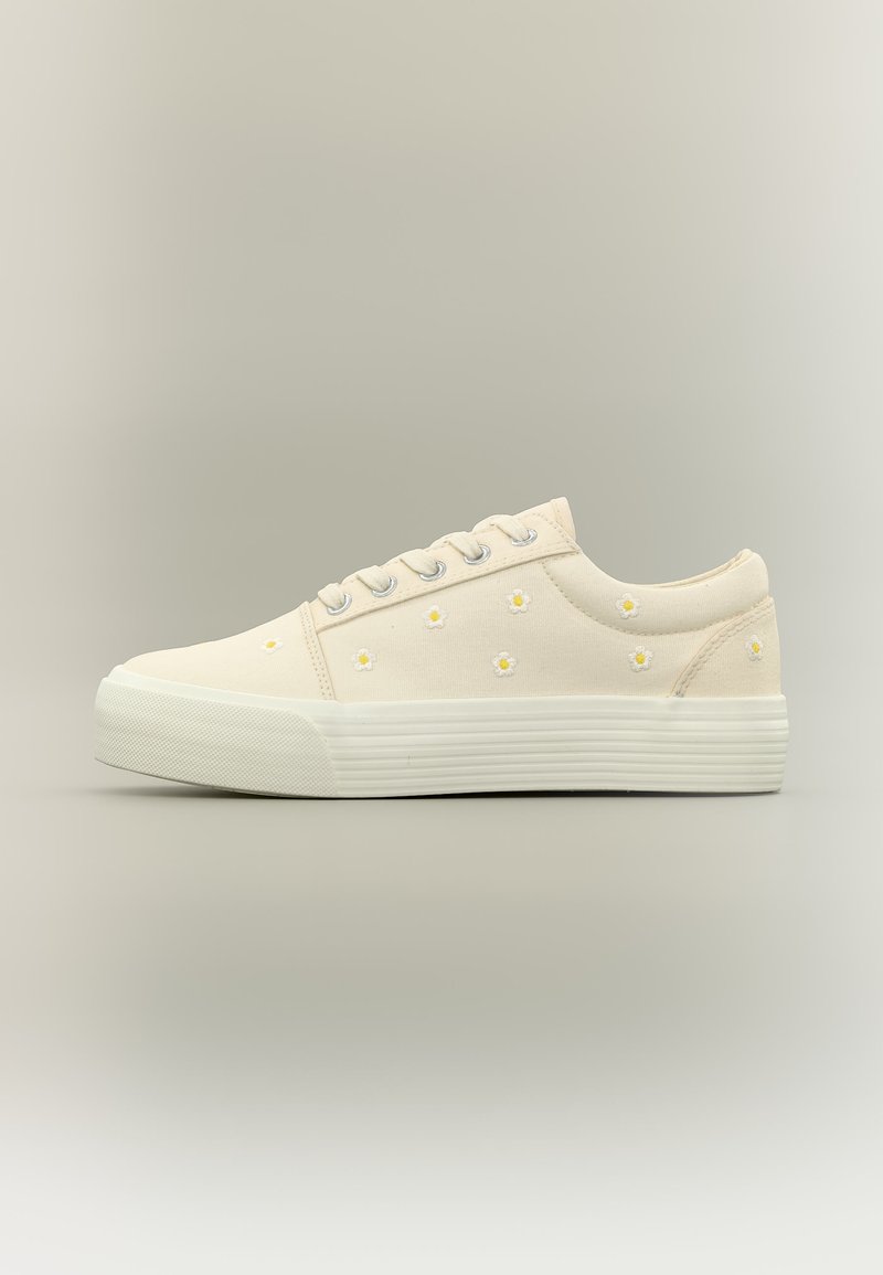 Κρεμ χαμηλή αθλητική παπούτσια με λευκή λαστιχένια σόλα, ασημί ρινίσματα και μικρά κεντημένα κίτρινα και λευκά μαργαρίτα πάνω στο υφασμάτινο πάνω μέρος.