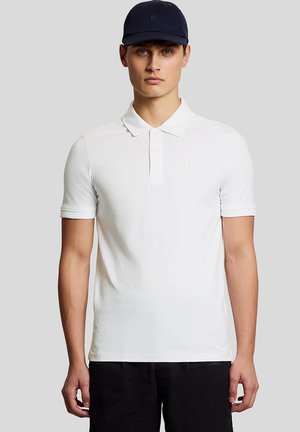 Jonge man die een wit poloshirt met korte mouwen en een marineblauwe pet draagt, staand tegen een effen lichtgrijze achtergrond.