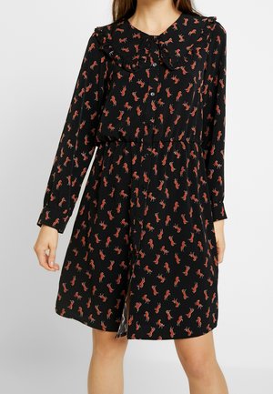 Czarna sukienka na długi rękaw z czerwonym nadrukiem konia, zapięciem na guziki z przodu, falbanianym kołnierzem i marszczonym stanem, noszona przez stojącą osobę.