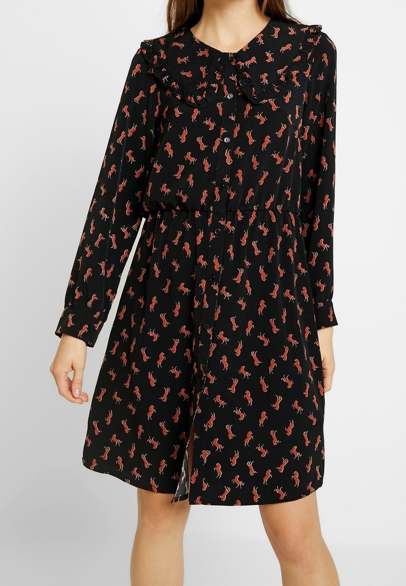 Robe noire à manches longues avec imprimé de chevaux rouges, devant boutonné, col à volants et taille froncée, portée par une personne debout.