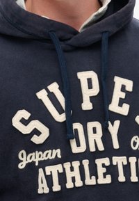 Close-up van een marineblauwe hoodie met witte geborduurde tekst: "Super Dry Japan Athletic" en blauwe trekkoorden.