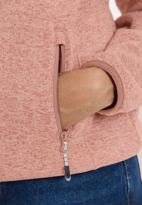 Rosa texturerad fleecejacka med en dragkedjeficka och figursydda ärmar. Har en kontrasterande dragkedjehake med ett mönstrat snöre.