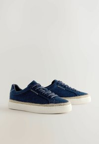 Blå jeansskor med vita gummisulor, kontrasterande sömmar och texturerade beige detaljer runt sulan. Snörningsdesign.