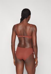 Bikini komplet v temno rjavo-rdeči barvi, ki ima nastavljive naramnice in bočne zanke na hlačkah. Tkanina z gladko teksturo, minimalističen dizajn.