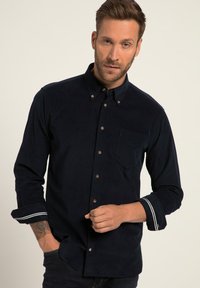 Camicia in corduroy blu navy con colletto a bottoni, taschino sul petto e maniche arrotolate con strisce bianche. Bottoni marroni aggiungono un tocco di dettaglio.