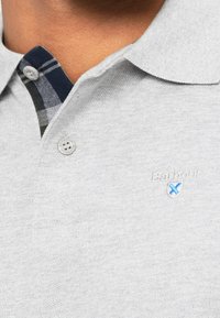 Polo gris clair avec deux boutons, laissant voir une couche intérieure à carreaux foncée et un petit logo brodé bleu et blanc sur la poitrine.