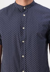 Camisa azul marino de manga corta con un patrón blanco, que cuenta con un cuello mao y botones beige en la parte delantera. Tejido ligero y diseño informal.