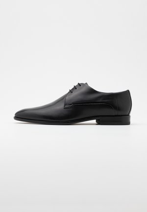 Chaussure de ville pour homme en cuir texturé noir, avec bout pointu et trois œillets pour lacets, sur un fond clair uni.